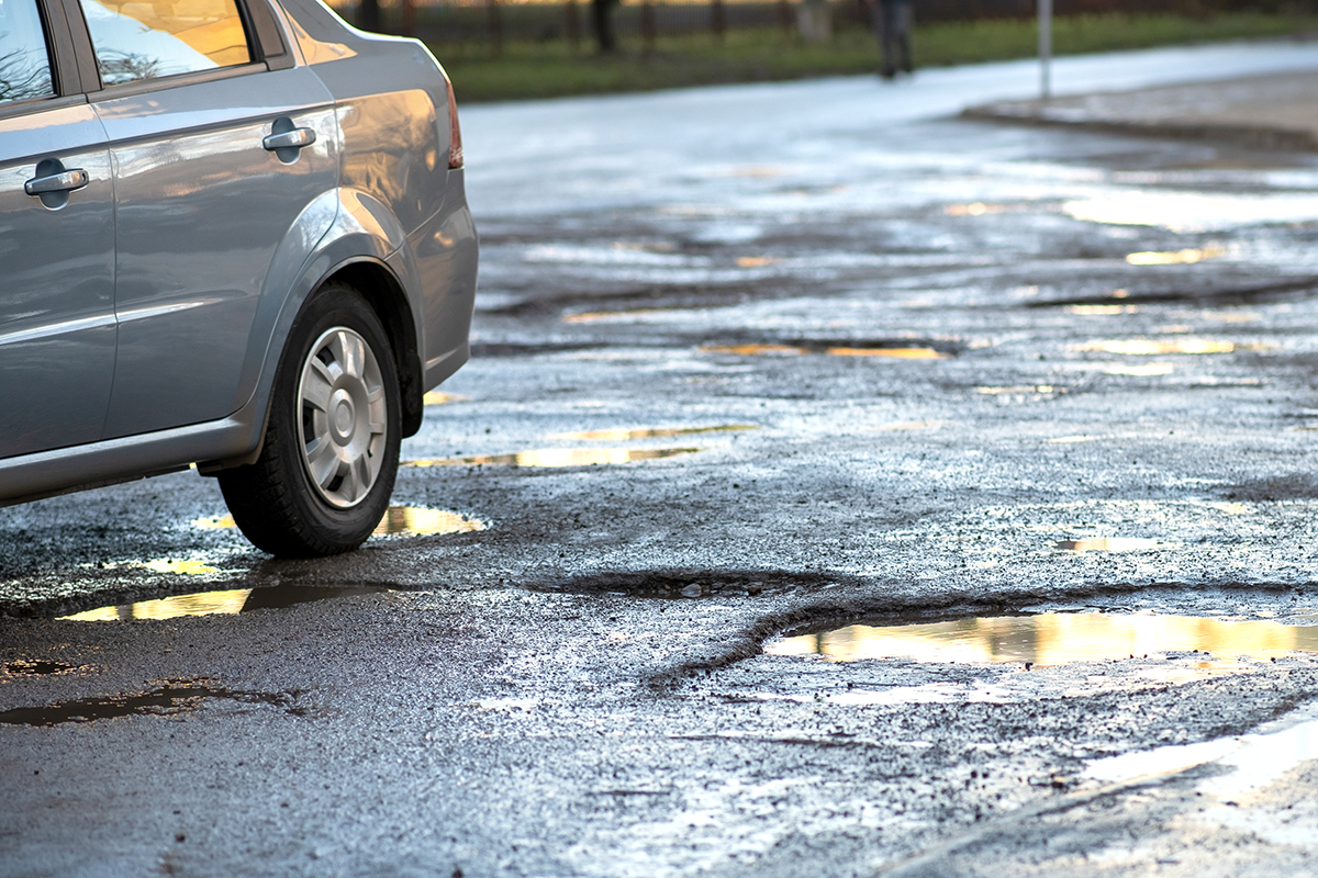 Car driving over potholes on a damaged road after winter, highlighting the importance of spring vehicle maintenance and suspension checks.